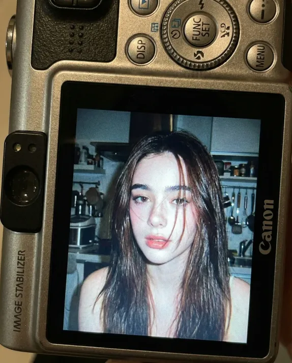 Use facial feature of attached photo. A close-up shot of a young woman displayed on the screen of a compact Canon digital camera. The camera body surrounds the image with its buttons, dials, and textured surface visible, including the FUNC/SET wheel, DISP button, and the "IMAGE STABILIZER" label along the side. The photo on the screen shows the woman indoors at night, illuminated by a bright built-in flash that creates sharp highlights on her face and hair. She has long dark hair falling across part of her face in loose strands, with a soft, slightly open-lip expression. The flash accentuates her features against a dim, cluttered kitchen background with appliances, shelves, and metallic surfaces softly blurred. The mood is candid, raw, nostalgic, and reminiscent of early 2000s digital camera snapshots. Colors are slightly muted with cool undertones, strong flash contrast, and natural grain from the display. No text, no logos inside the photo preview itself.
Camera: compact digital camera simulation
Lens: equivalent to 28-35mm
Aperture: f/2.8
ISO: 400
Shutter speed: 1/60 with flash
White balance: auto flash
Lighting: harsh direct flash on subject, ambient low light in the background
Color grading: nostalgic digital-camera tones, high contrast flash, subtle display grain, authentic screen glow.